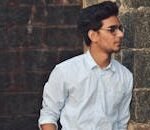 A stylish young man in glasses leans against a brick wall wearing a casual light blue shirt.
